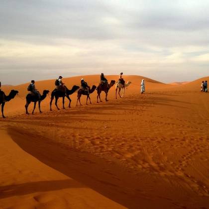 A Group Traveling On Camels Over Reddish Sands Under A Cloudy Sky In A Vast Desert Landscape.