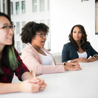 Three Diverse Women Discussing In A Bright Office, Highlighting Teamwork And Collaboration.