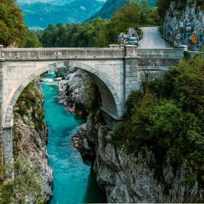 Scenic View Of A Stone Arch Bridge Over A Vibrant Turquoise River Framed By Lush Greenery.