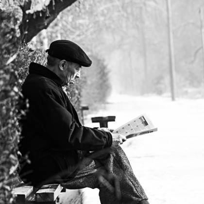 A Senior Man Reads A Newspaper On A Snowy Bench In A Tranquil Winter Park.