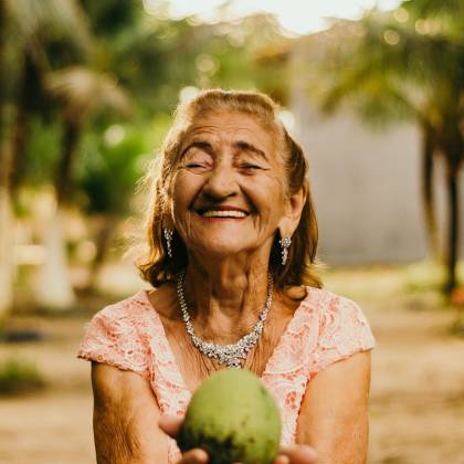 Smiling Elderly Woman With Jewelry Showcases Happiness While Holding A Mango Outdoors.