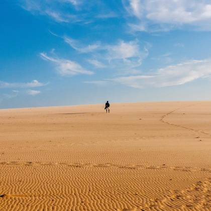 A Lone Traveler Walks Across A Vast And Serene Desert Under A Bright Blue Sky.
