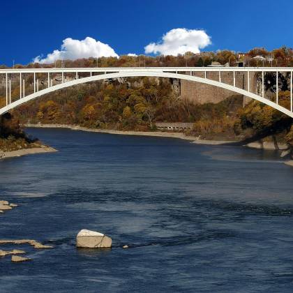 Panoramic View Of Rainbow Bridge Over The River In Fall With Vibrant Foliage And Blue Sky.