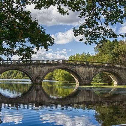 Ancient Stone Bridge Arches Over A Calm River, Surrounded By Lush Greenery And Clear Skies.