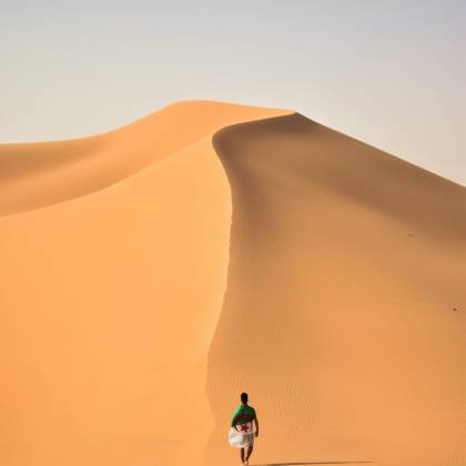 A Lone Traveler Walking Over The Vast, Golden Sand Dunes Of The Sahara Desert In Algeria.