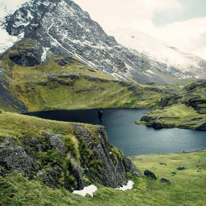 A Hiker Stands On A Cliff Overlooking A Serene Mountain Lake Surrounded By Lush Greenery And Snow Capped Peaks.