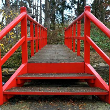 A Vibrant Red Wooden Bridge Crossing A Serene Stream In A Tranquil Autumn Park.