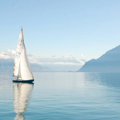 Sailing Boat Glides Peacefully On A Misty Lake Surrounded By Mountains, Under Clear Daylight.