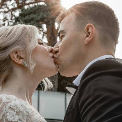 Couple Sharing A Romantic Kiss On Their Wedding Day With Sunshine Peeking Through Trees.