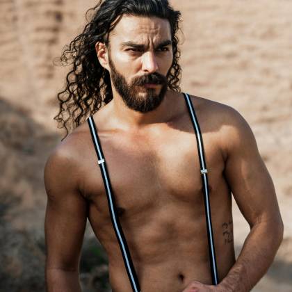 Shirtless Man With Curly Hair And Suspenders Posing Confidently Outdoors.
