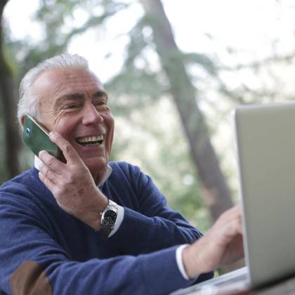 Happy Senior Man Talking On Phone And Using Laptop Outdoors.