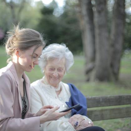 Side View Of Smiling Adult Female Helping Aged Mom In Using Of Mobile Phone While Sitting Together In Park