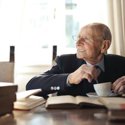 Positive Senior Man In Formal Suit And Eyeglasses Drinking Hot Beverage From Cup While Sitting At Wooden Table With Books And Looking Away