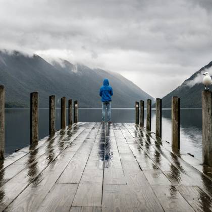 Person In Blue Jacket Stands On A Rainy Dock Overlooking Serene Lake And Mountains.