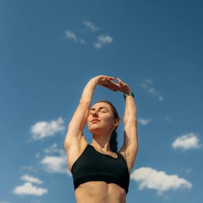 A Woman In Black Leggings And Sports Bra Stretches Under A Clear Blue Sky, Promoting Fitness And Freedom.